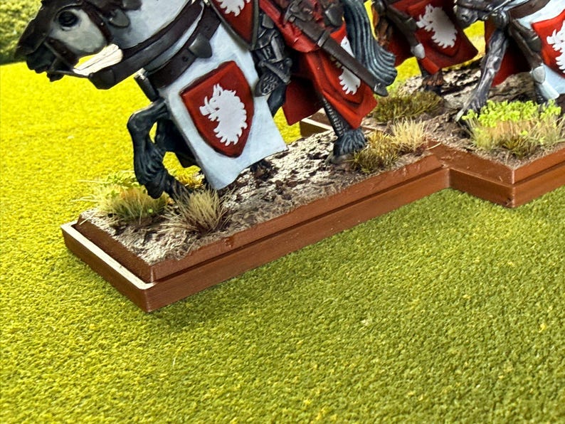 Peut inclure: Figurines miniatures de chevaliers &agrave; cheval. Les chevaux sont blancs et noirs, avec une armure d&eacute;taill&eacute;e et des &eacute;cussons h&eacute;raldiques rouges et blancs. La base est marron avec de l'herbe et de la terre factices, pos&eacute;e sur une surface verte.
