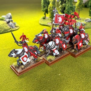 Peut inclure: Figurines miniatures de chevaliers &agrave; cheval, peintes en rouge et blanc, avec des boucliers et des banni&egrave;res. Les chevaliers sont pos&eacute;s sur une surface herbeuse verte, avec des arbres en arri&egrave;re-plan. Les figurines sont probablement pour un jeu de soci&eacute;t&eacute;.