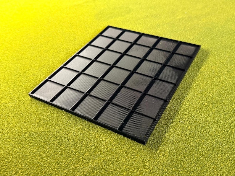 May include: A black, rectangular tray with a grid pattern of square compartments. The tray is made of a matte black material and sits on a textured, yellow-green surface. The grid is evenly spaced, creating a series of small squares.