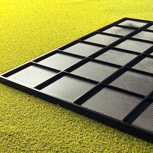 May include: A black tray with a grid pattern, featuring numerous square compartments. The tray has a textured surface and is set against a textured, yellow-green background. The compartments are divided by raised black lines.