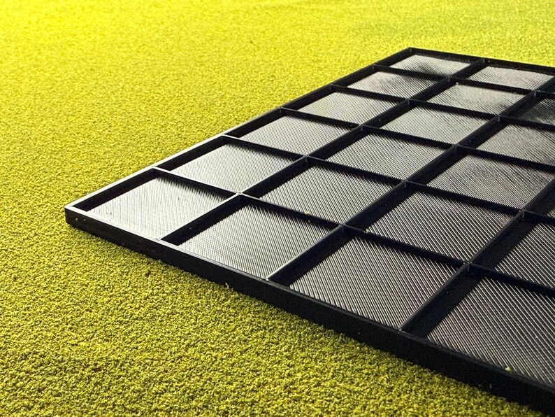 May include: A black tray with a grid pattern, featuring numerous square compartments. The tray has a textured surface and is set against a textured, yellow-green background. The compartments are divided by raised black lines.