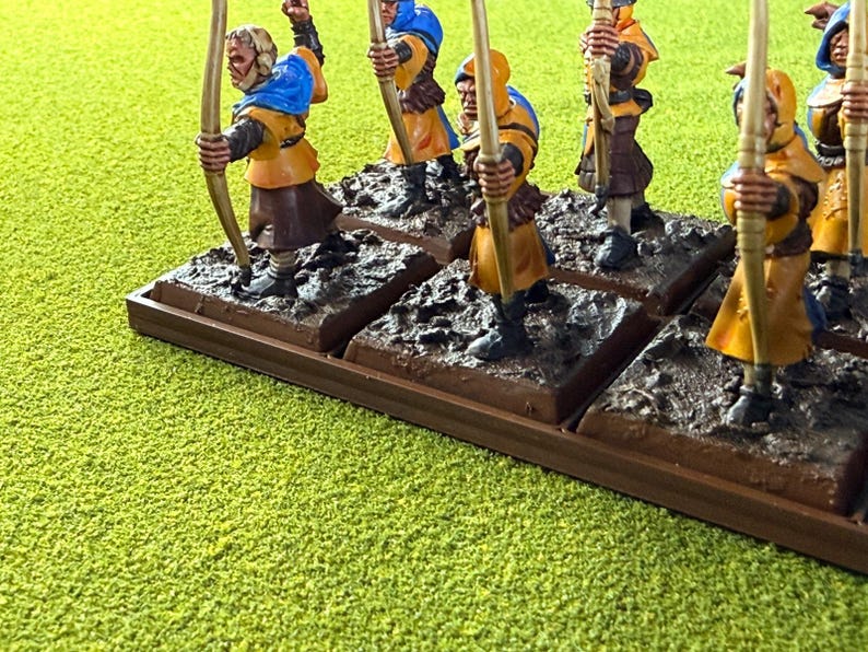 May include: Miniature archer figurines in yellow and blue, each with a bow and arrow. The figures are on individual bases, mounted on a larger brown base. The scene is set against a green, textured background.
