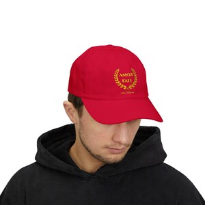 Könnte beinhalten: Eine leuchtend rote Baseballkappe mit einem goldfarbenen, gestickten Lorbeerkranz auf der Vorderseite. Innerhalb des Kranzes steht der Text AMOR FATI in goldenen Großbuchstaben. Die Kappe hat eine strukturierte Krone und einen gebogenen Schirm.