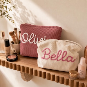 Puede incluir: Dos neceseres de maquillaje de pana, uno rosa empolvado con "Olivia" en escritura blanca y otro crema con "Bella" en escritura rosa. Los neceseres están sobre una estantería de madera con brochas de maquillaje y productos para el cuidado de la piel.