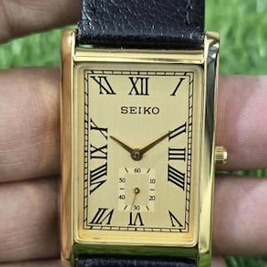May include: A rectangular Seiko wristwatch featuring a gold-tone dial with black Roman numeral hour markers and a sub-dial for seconds. The watch has a polished gold-tone case and a textured black leather strap with a buckle closure.