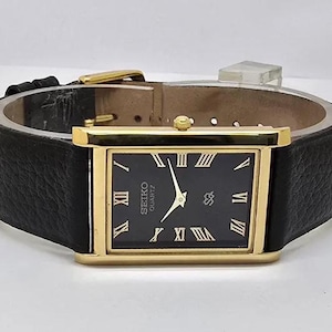 Peut inclure: Une montre-bracelet rectangulaire Seiko Quartz avec un boîtier doré, un cadran noir doté de chiffres romains dorés et des aiguilles dorées. La montre est fixée à un bracelet en cuir noir texturé avec une boucle dorée et une couronne latérale.