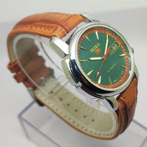 May include: A Seiko 5 automatic wristwatch featuring a round silver-tone stainless steel case, a textured green dial with gold-tone indices, a day-date window, and a light brown leather strap with a crocodile-embossed pattern and white stitching.