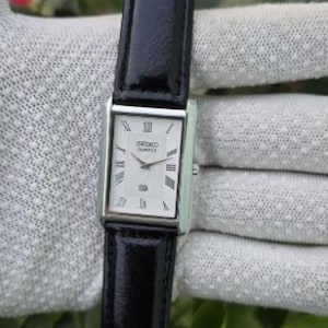 May include: A rectangular Seiko Quartz wristwatch featuring a white dial with black Roman numeral hour markers and silver-tone hands. The watch has a polished silver-tone case and a glossy black leather strap with a matching black buckle.