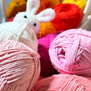 May include: A white crocheted bunny sits among a basket of colorful yarn balls. The yarn is in shades of pink, red, orange, and yellow. The bunny has black eyes and pink inner ears. The yarn is soft and fluffy, perfect for crafting.