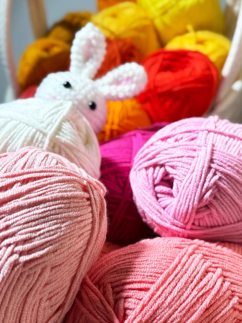 May include: A white crocheted bunny sits among a basket of colorful yarn balls. The yarn is in shades of pink, red, orange, and yellow. The bunny has black eyes and pink inner ears. The yarn is soft and fluffy, perfect for crafting.