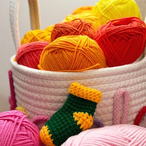 May include: A white woven basket filled with colorful yarn balls in shades of yellow, orange, and red. A small, green and yellow crocheted sock sits in front of the basket, with pink yarn balls in the foreground.