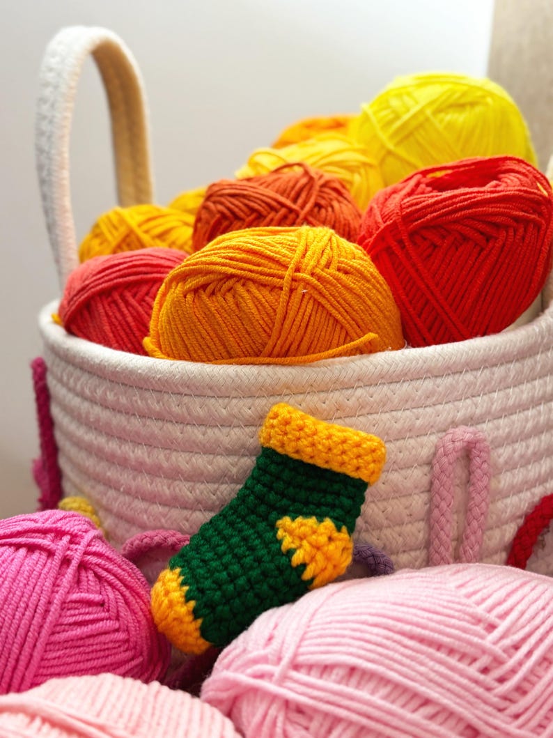May include: A white woven basket filled with colorful yarn balls in shades of yellow, orange, and red. A small, green and yellow crocheted sock sits in front of the basket, with pink yarn balls in the foreground.