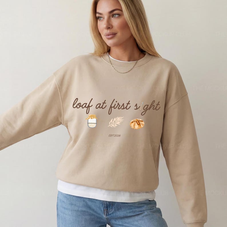 May include: Beige sweatshirt with the phrase "loaf at first sight" in brown script. The design includes watercolor illustrations of a jar, wheat, and a loaf of bread. The text "EST 2026" is below the wheat illustration.
