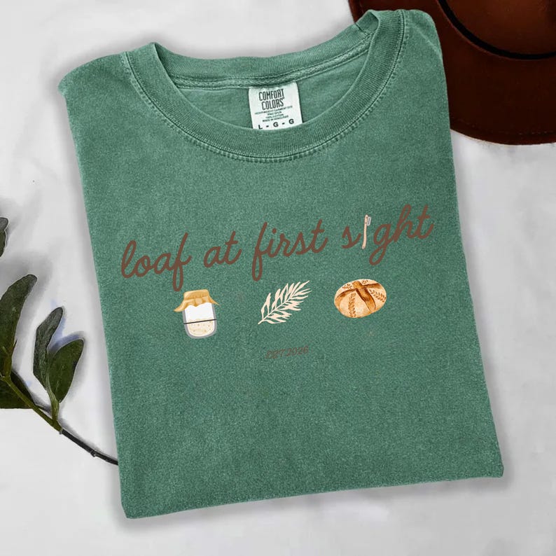 May include: Green Comfort Colors t-shirt with the phrase "loaf at first sight" in brown script. The shirt features watercolor illustrations of a jar, a sprig, and a loaf of bread. A brown hat is visible in the background.