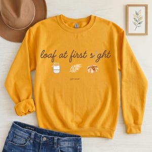May include: Mustard yellow sweatshirt with the phrase "loaf at first sight" in a script font. The design includes illustrations of a jar, a sprig, and a loaf of bread. The text "EST.2026" is below the illustrations.