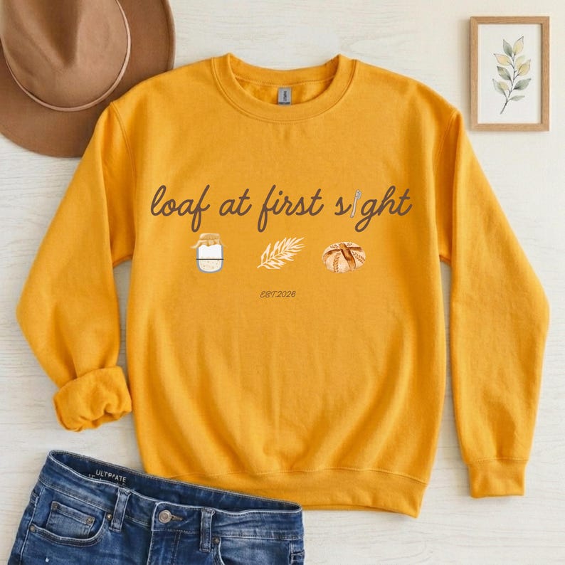 May include: Mustard yellow sweatshirt with the phrase "loaf at first sight" in a script font. The design includes illustrations of a jar, a sprig, and a loaf of bread. The text "EST.2026" is below the illustrations.