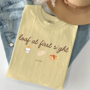 May include: A light yellow t-shirt with the phrase "loaf at first sight" in brown script. The shirt features illustrations of a jar of jam, a sprig, and a loaf of bread. The text "EST 2026" is also printed on the shirt.