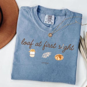 May include: A blue Comfort Colors t-shirt with the phrase "loaf at first sight" in brown script. The shirt features illustrations of a jar of jam, a sprig, and a loaf of bread. A gold necklace with a pendant is draped across the shirt.