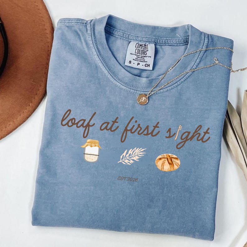 May include: A blue Comfort Colors t-shirt with the phrase "loaf at first sight" in brown script. The shirt features illustrations of a jar of jam, a sprig, and a loaf of bread. A gold necklace with a pendant is draped across the shirt.