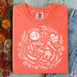 Może przedstawiać: Koralowa koszulka z białym okrągłym nadrukiem przedstawiającym ilustracje związane z pieczeniem i napisem "ARTISANAL". Projekt zawiera bochenek chleba, miarkę, trzepaczkę i inne przybory kuchenne. Koszulka jest złożona na drewnianej powierzchni.