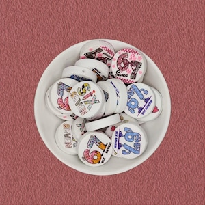 May include: A collection of round white beads featuring various colorful graphic designs of the number 67 and the text "Six Seven". The beads are displayed in a white bowl, showcasing multiple artistic styles including polka dots, stripes, and cartoon characters.