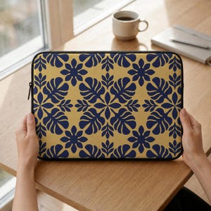 May include: A laptop sleeve with a navy blue floral pattern on a gold background. The sleeve has a black zipper and is held by a person. A coffee cup and notebook are in the background.