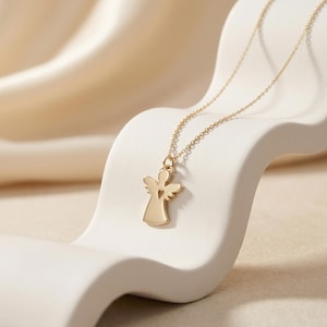 Puede incluir: Un collar con colgante de &aacute;ngel dorado. El colgante presenta la silueta de un &aacute;ngel con un coraz&oacute;n recortado. La cadena del collar es delicada y dorada. El collar se exhibe sobre una superficie blanca y curvada.