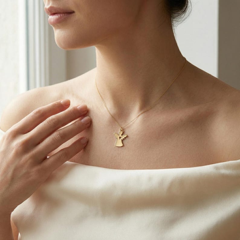 Puede incluir: Un collar con colgante de &aacute;ngel dorado. El colgante presenta una silueta de &aacute;ngel estilizada con alas extendidas y un recorte en forma de coraz&oacute;n. El collar se lleva en el cuello de una persona.