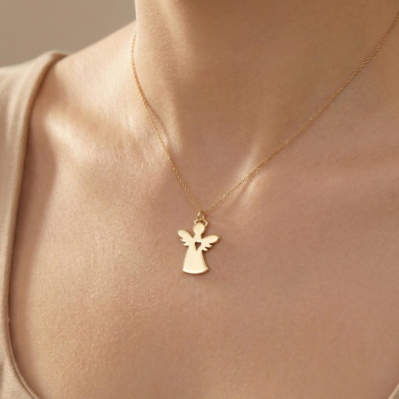 Puede incluir: Un collar con colgante de &aacute;ngel dorado. El colgante presenta una silueta de &aacute;ngel con un coraz&oacute;n recortado. El collar se lleva alrededor del cuello, con la cadena y el colgante visibles.