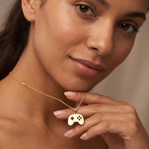 Può includere: Una collana color oro con un ciondolo a forma di controller per videogiochi. Il ciondolo presenta un design traforato con una croce direzionale a sinistra e quattro pulsanti circolari a destra, sospeso a una sottile catenina dorata.