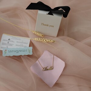 Puede incluir: Un collar dorado con el nombre "Bella" sobre una tela rosa, presentado con una caja de regalo y un lazo negro. La caja tiene la inscripci&oacute;n "Thank you". Tambi&eacute;n se incluyen espigas de trigo decorativas y una peque&ntilde;a tarjeta.