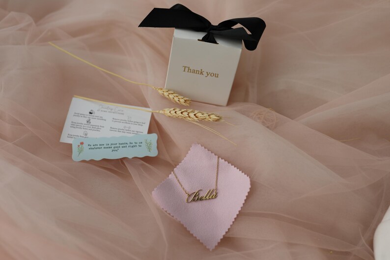 Puede incluir: Un collar dorado con el nombre "Bella" sobre una tela rosa, presentado con una caja de regalo y un lazo negro. La caja tiene la inscripci&oacute;n "Thank you". Tambi&eacute;n se incluyen espigas de trigo decorativas y una peque&ntilde;a tarjeta.