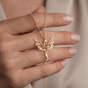 May include: A gold-colored pendant necklace featuring a stylized phoenix design. The pendant is a delicate outline of the mythical bird with outstretched wings. The necklace is held by a hand, showcasing the intricate details of the jewelry.