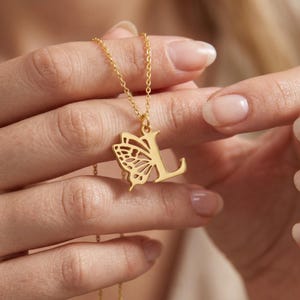 Puede incluir: Un collar dorado con un colgante en forma de la letra "L" y un ala de mariposa. El ala de mariposa está en el lado izquierdo de la letra. El collar está en una cadena delicada.