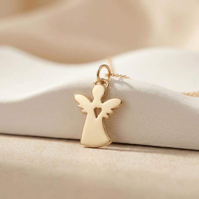 Puede incluir: Un collar con colgante de &aacute;ngel dorado. El colgante tiene forma de &aacute;ngel con un recorte en forma de coraz&oacute;n. Las alas est&aacute;n extendidas y el collar est&aacute; en una cadena dorada. El fondo es de color neutro.