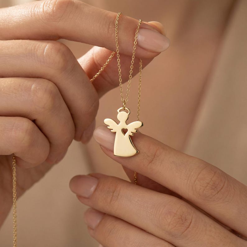 Puede incluir: Un collar con colgante de &aacute;ngel dorado. El colgante presenta la silueta de un &aacute;ngel con un coraz&oacute;n recortado en el centro. El collar est&aacute; sujeto por una persona. La cadena es fina y el colgante tiene un acabado liso.