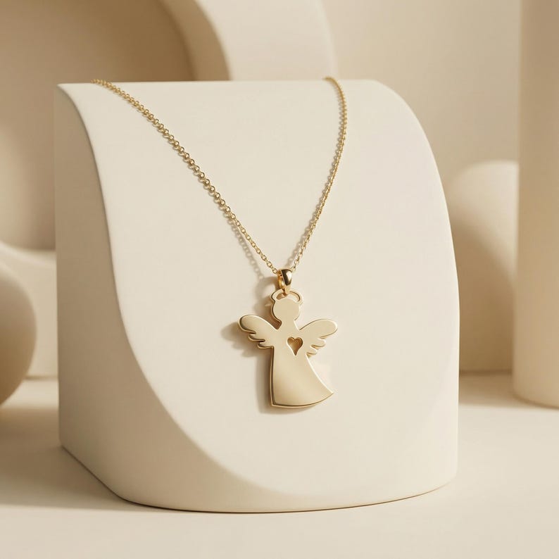 Puede incluir: Un collar con colgante de &aacute;ngel dorado. El colgante presenta una silueta de &aacute;ngel con un coraz&oacute;n recortado. El collar se exhibe sobre un pedestal blanco curvado sobre un fondo neutro. La cadena es delicada y dorada.