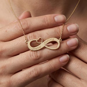 Puede incluir: Un collar infinito dorado con los nombres "Joanne" y "Anthony" grabados. El collar presenta un dije en forma de corazón y está suspendido de una delicada cadena. La joya se exhibe sobre un fondo neutro.