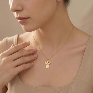 Puede incluir: Un collar con colgante de &aacute;ngel dorado. El colgante presenta una silueta de &aacute;ngel con un recorte en forma de coraz&oacute;n. El collar se lleva alrededor del cuello de una persona.