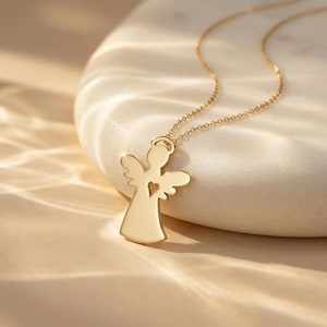 Puede incluir: Un collar con colgante de &aacute;ngel dorado. El colgante presenta la silueta de un &aacute;ngel con un coraz&oacute;n recortado. El collar se muestra sobre una superficie de piedra blanca, con una iluminaci&oacute;n suave que crea sombras.