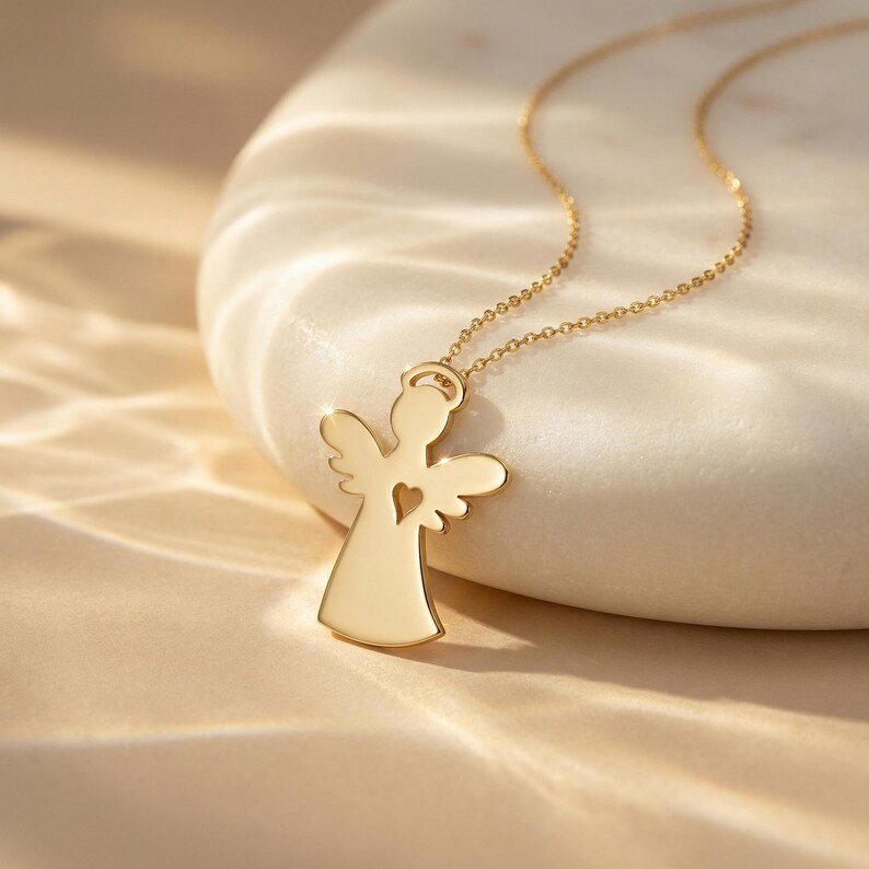 Puede incluir: Un collar con colgante de &aacute;ngel dorado. El colgante presenta la silueta de un &aacute;ngel con un coraz&oacute;n recortado. El collar se muestra sobre una superficie de piedra blanca, con una iluminaci&oacute;n suave que crea sombras.