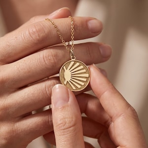 May include: A gold-toned circular pendant necklace featuring a textured background and a raised, stylized sunburst or shell-like motif with radiating lines. The pendant is suspended from a delicate, matching gold-toned metal link chain.