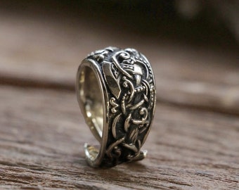 May include: A silver ring with an intricate design featuring a knotwork pattern and a stylized animal figure. The ring is shown on a wooden surface.