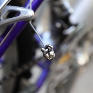 May include: A small, metallic, anatomically shaped heart charm suspended by a thin wire from the frame of a purple bicycle. The charm features a polished, silver-toned finish with visible details of the heart's chambers and major blood vessels.