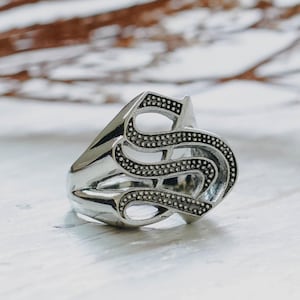 Puede incluir: Un anillo de metal en tono plateado que presenta una letra S estilizada y prominente como pieza central. La letra tiene un acabado de superficie texturizado y punteado dentro de sus curvas, mientras que la banda y los bordes permanecen lisos.