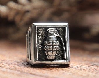 May include: A silver-colored ring with a rectangular face featuring a detailed grenade design. The ring has a polished finish and a bold, masculine aesthetic. The grenade is depicted in a three-dimensional style, adding depth to the jewelry.