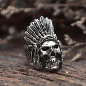 May include: A silver-colored ring featuring a skull wearing a Native American headdress. The ring has intricate details, including feathers and facial features. The ring is set against a wooden surface.