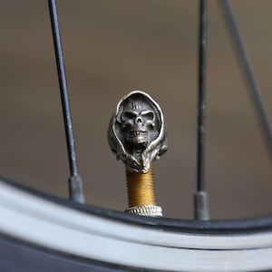 May include: A silver-toned metal valve cap shaped like a hooded skull, attached to the threaded brass valve stem of a bicycle tyre. The skull features detailed eye sockets, a nose cavity, and teeth, with a dark, weathered finish on the metal surface.