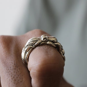 Könnte beinhalten: Ein goldfarbener Ring mit einem Oktopus-Design. Der Ring ist mit strukturierten Tentakeln und einem zentralen Kopf versehen, die ein maritimes Thema zeigen. Der Ring wird an einem Finger getragen.