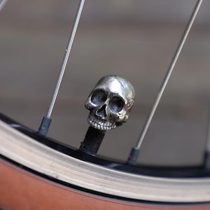 May include: A metallic silver-colored skull-shaped valve cap attached to a bicycle tire valve stem. The skull features detailed eye sockets, a nasal cavity, and teeth, positioned between the thin metal spokes of a bicycle wheel rim.